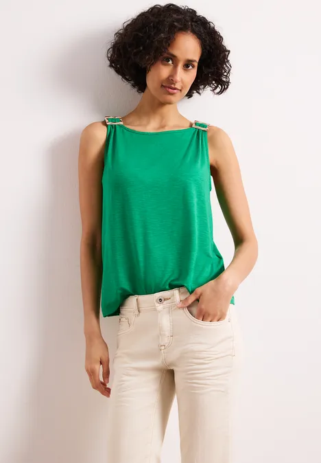 Frau mit lockigem Haar trägt ärmelloses grünes Top mit Zierträgern und beigefarbene Hose.