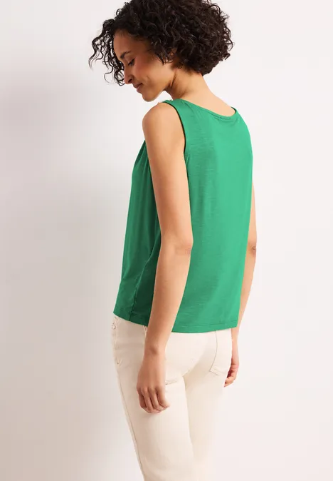 Frau von hinten in grünem ärmellosem Top und beigen Hosen vor hellem Hintergrund.