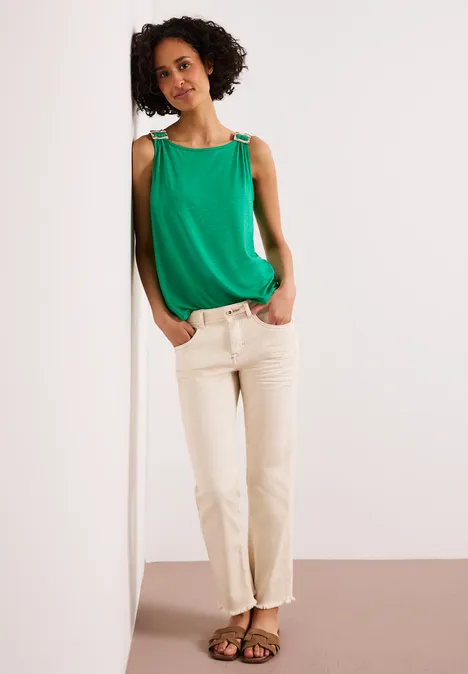 Eine Person lehnt an einer weißen Wand, trägt grünes Top, beige Hose und Sandalen.