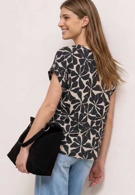 Vrouw met lang blond haar van achteren gezien, draagt een zwart-wit bloemmotief T-shirt, blauwe jeans en zwarte tas.