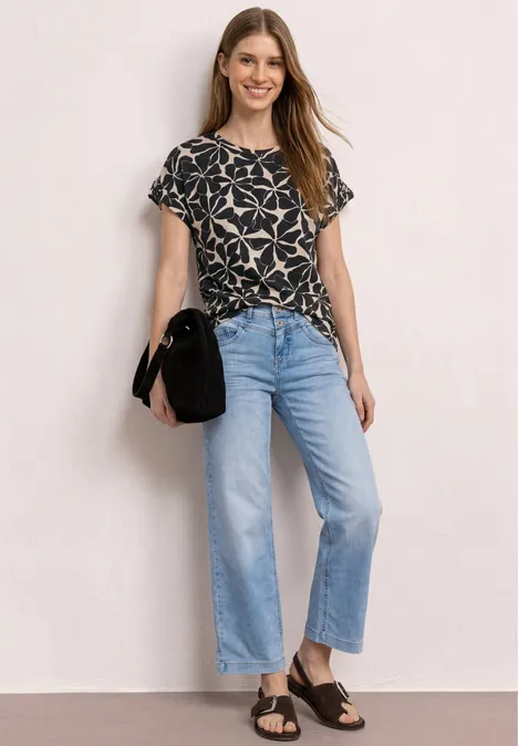 Vrouw in zwart patroon-T-shirt, lichtblauwe jeans, zwarte sandalen en zwarte tas tegen een lichte achtergrond.