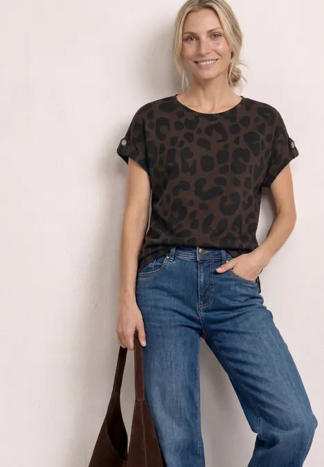 Frau in lässigem Outfit: Leo-T-Shirt, blaue Jeans und braune Tasche vor weißem Hintergrund.