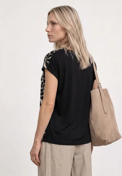 Eine blonde Frau in schwarzem T-Shirt und beigen Hosen mit großer beiger Tragetasche, Rückenansicht.
