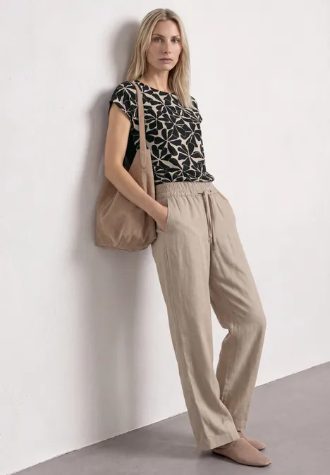 Frau lehnt an Wand, trägt beige Leinenhose, gemustertes Top, große Tasche