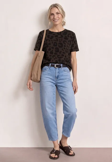 Persoon voor witte muur draagt zwart leopard-T-shirt, lichte jeans, zwarte sandalen en katoenen tas.