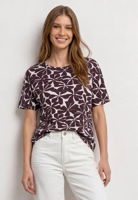 Vrouw in paars-wit t-shirt met patroon en lichte broek voor een neutrale achtergrond.