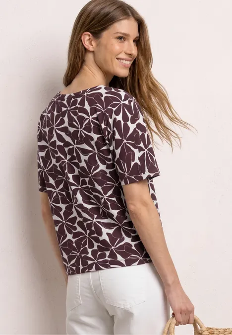 Vrouw van achteren in bruin-witte bladprint top, witte broek en met een strooien tas.