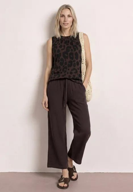 Top à col rond et imprimé zèbre molten earth Frau trägt ärmelloses Leoparden-Top, weite schwarze Hose, Sandalen und Basttasche.