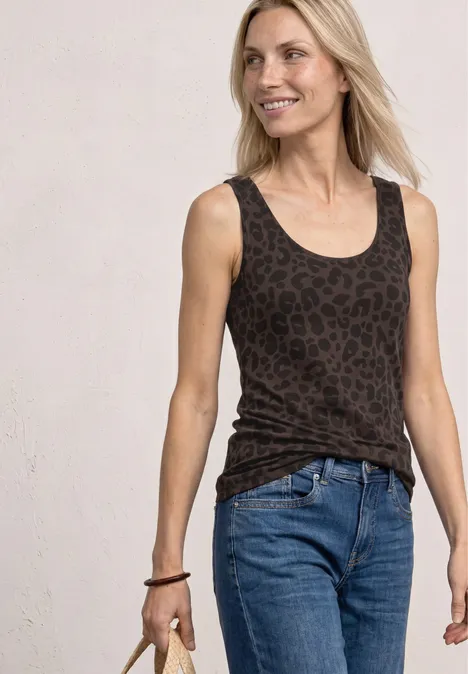 Eine Frau in Jeans und dunklem, leo-bedrucktem Tanktop vor hellem Hintergrund.