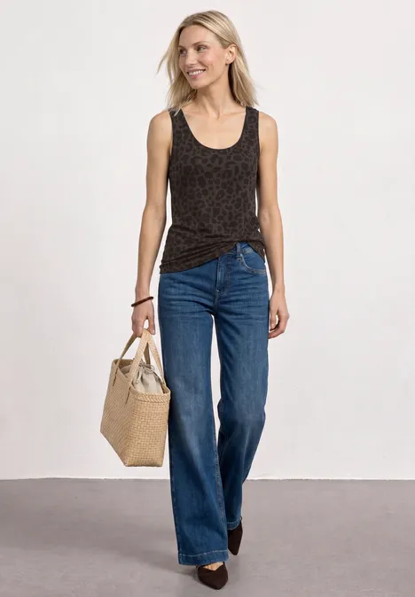 Eine Frau trägt ein ärmelloses, schwarzes Top mit Muster, weite blaue Jeans und hält eine Strohtasche.