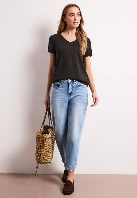 Frau in schwarzem V-Neck-Shirt, lockeren blauen Jeans, braunen Loafers, trägt einen geflochtenen Korb als Tasche.