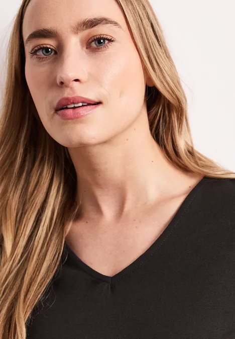 Frau mit langem Haar in schwarzem V-Ausschnitt-Shirt vor hellem Hintergrund.