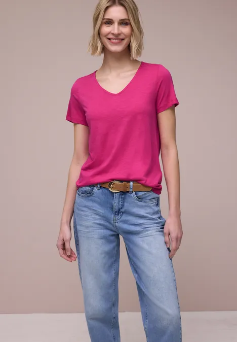 Persoon in roze T-shirt, blauwe jeans en bruine riem tegen neutrale achtergrond.