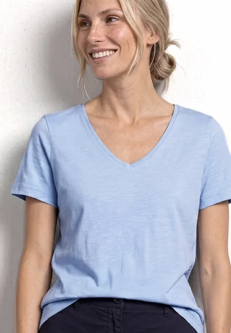 Eine lächelnde Frau in hellblauem V-Ausschnitt-Shirt und dunkler Hose vor hellem Hintergrund.