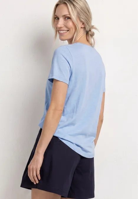 Frau steht seitlich, trägt hellblaues T-Shirt und schwarze Shorts vor weißem Hintergrund.