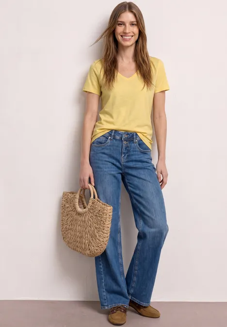 Femme en t-shirt jaune et jean large, tenant un sac en paille, sur fond clair.