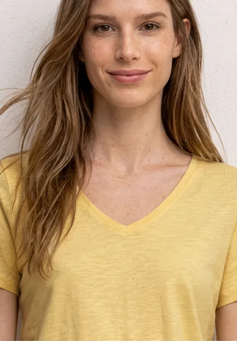 Femme en t-shirt jaune à col V, cheveux longs, devant un mur blanc.