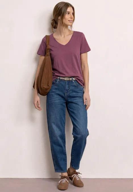 Vrouw leunt tegen muur, draagt paars T-shirt, blauwe high-waist jeans, bruine schoenen en tas.