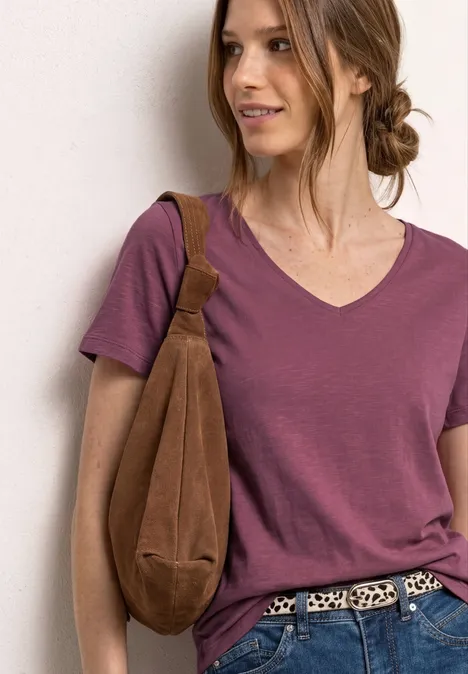 Vrouw met opgestoken haar, paarse V-hals T-shirt, luipaardriem en bruine schoudertas voor lichte achtergrond.