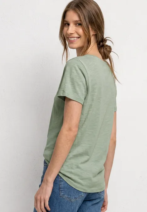 Seitlich abgebildete Frau mit grünem T-Shirt und blauen Jeans vor einem hellen Hintergrund.