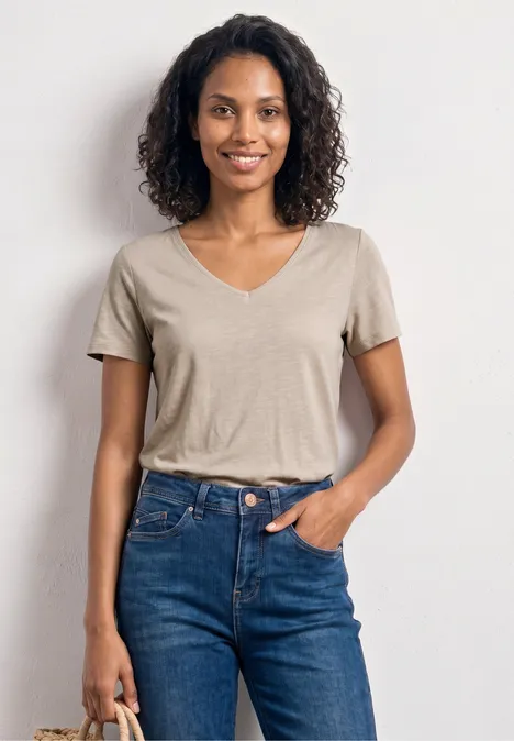 Frau in beigem V-Neck-Shirt und blauen Jeans steht vor weißem Hintergrund und hält Tasche.