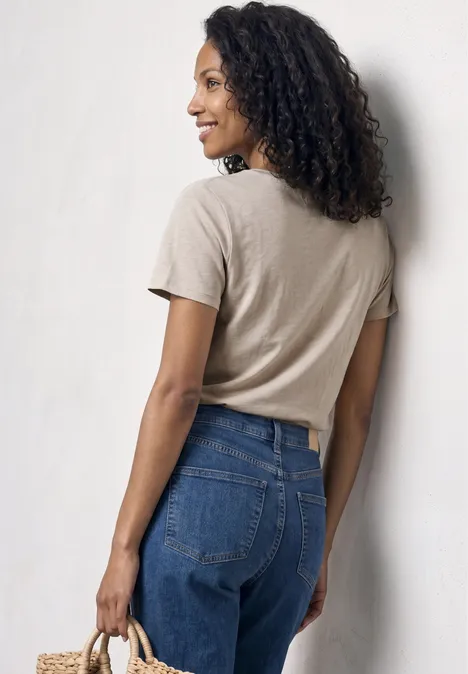 Frau von hinten in beigefarbenem T-Shirt, blauen Jeans und Strohtasche in der Hand.