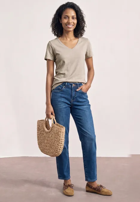 Frau mit lockigem Haar in beigem T-Shirt, blauen Jeans und braunen Loafers hält eine Strohtasche