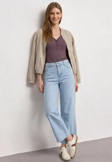 Top à col en V et à rayures rustic rouge Une femme pose devant un mur blanc, porte un jean droit bleu clair, un tee-shirt rayé et une veste beige.