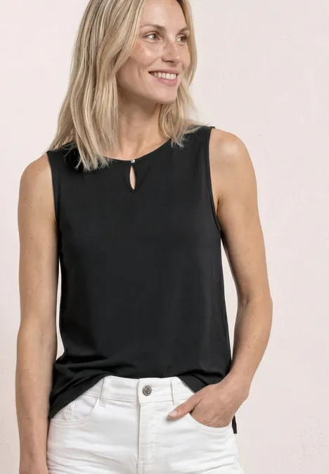 Femme portant un haut noir sans manches et un pantalon clair, main dans la poche, fond lumineux.