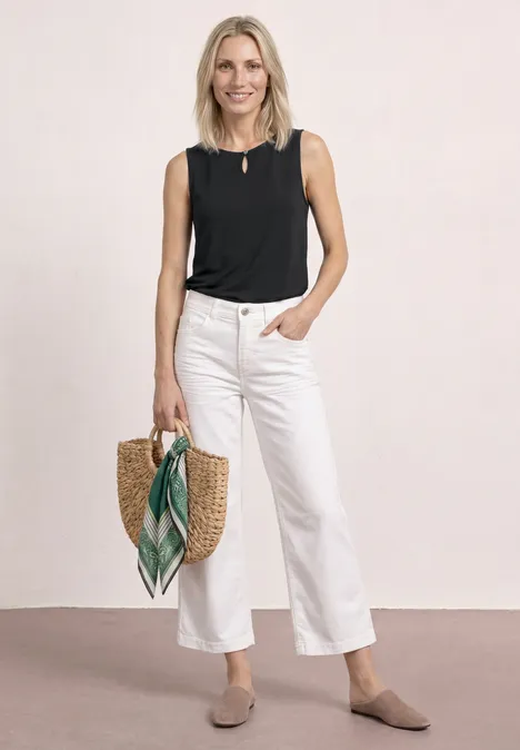 Femme en top noir sans manches et pantalon blanc large, mocassins bruns, portant un sac en paille avec foulard