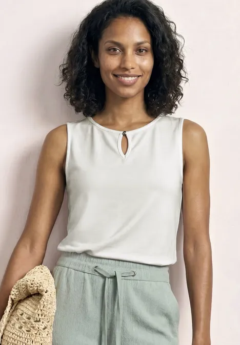 Femme en haut blanc sans manches et pantalon vert clair, tenant un sac en paille.
