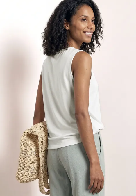 Femme vu de dos, cheveux bouclés, porte un débardeur blanc, un pantalon gris et un sac tressé beige.