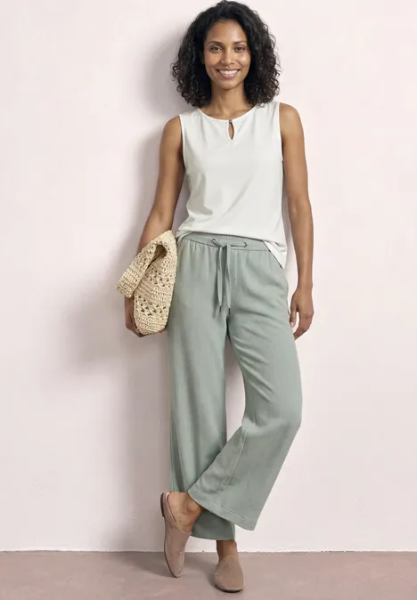 Femme en débardeur blanc et pantalon ample vert avec sac en paille, pieds nus devant un mur clair.