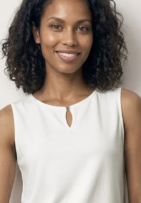 Femme en top blanc sans manches à découpe clé, cheveux bouclés foncés, visage flou.