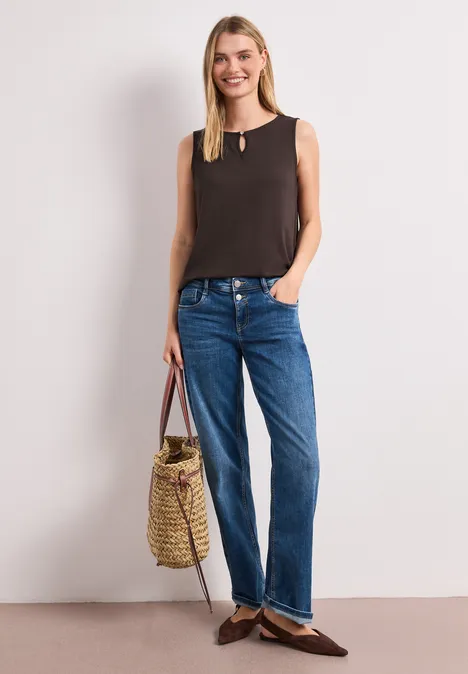Femme vêtue d’un débardeur noir, jean bleu et mocassins bruns, tenant un sac en paille, posant devant un fond blanc.