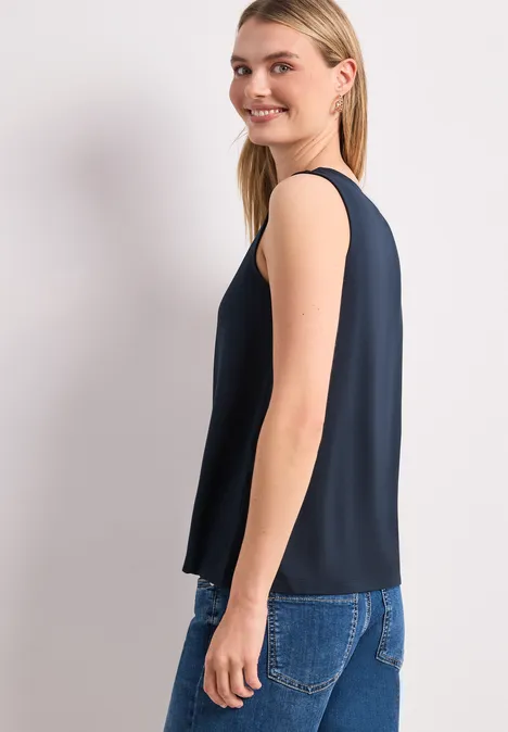 Une femme en top sans manches bleu foncé et jean, vue de profil sur fond clair.