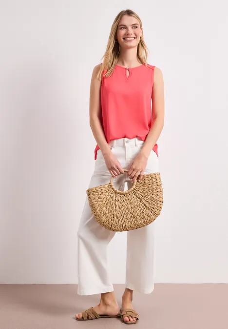 Vrouw in roze top en wijd witte broek, met sandalen en strooien tas voor een neutrale achtergrond.