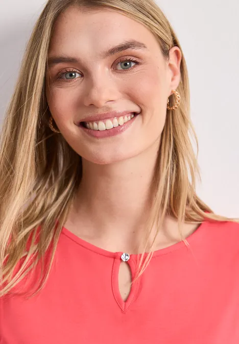 Vrouw in een koraalroze top met druppeluitsnijding, lang blond haar en subtiele oorbellen.
