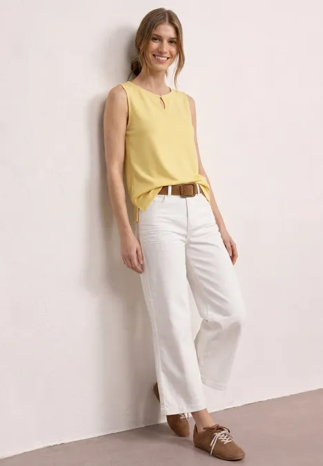 Person in gelbem Top, weißen Hosen und braunen Schuhen lehnt an heller Wand.