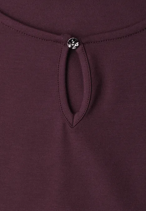 Haut bordeaux avec petite ouverture en forme de goutte au col, fermé par un bouton rond décoratif.