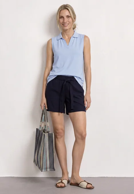 Top in Rippstruktur mit Polokragen cashmere blue Frau in hellblauem ärmellosem Polo, dunkelblauen Shorts, gestreifter Tasche und Sandalen vor weißem Hintergrund.