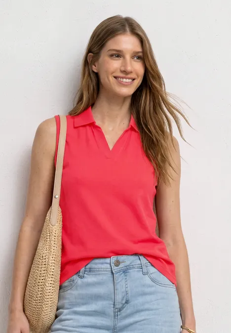 Vrouw tegen witte muur in rood mouwloos poloshirt, blauwe jeans en rieten tas.