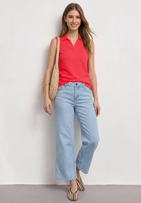 Vrouw in rode mouwloze polo, lichtblauwe wijde jeans, gestreepte sandalen en strozak voor een witte muur.