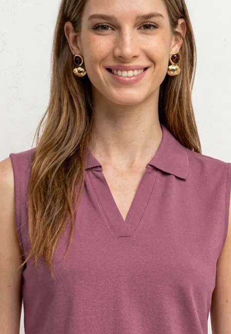 Een vrouw met lang bruin haar draagt een mouwloze paarse polo en gouden creolen.