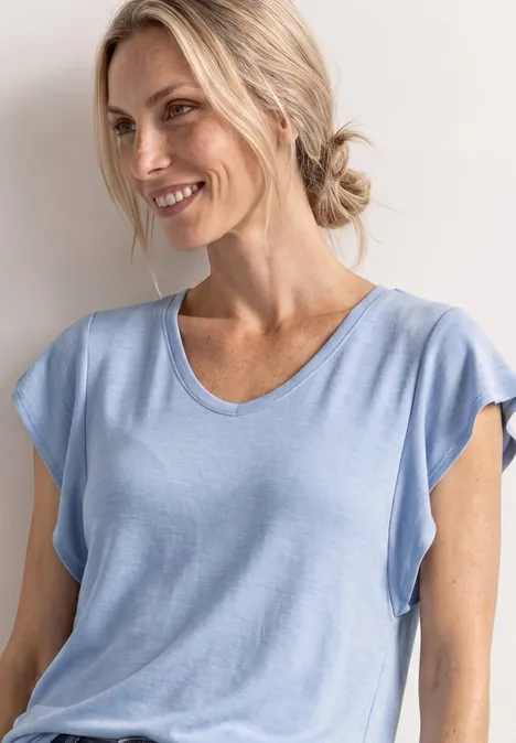 Eine Frau mit blondem Haar zu einem Dutt gebunden trägt ein hellblaues T-Shirt und schaut seitlich.
