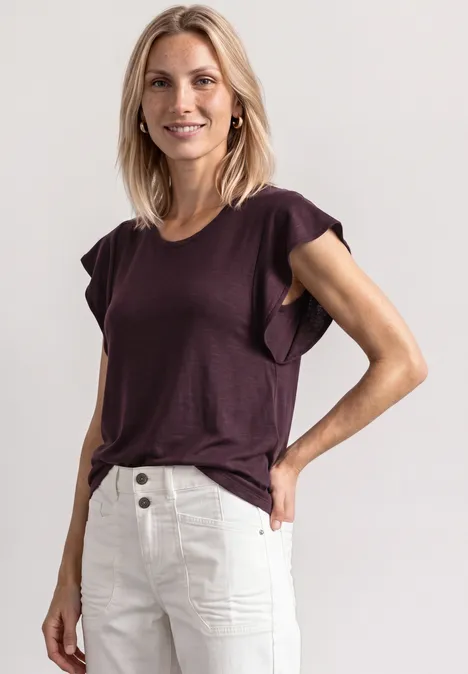 Eine Frau in einem violetten T-Shirt mit Rüschenärmeln und hoch geschnittenen cremefarbenen Hosen in lässiger Pose