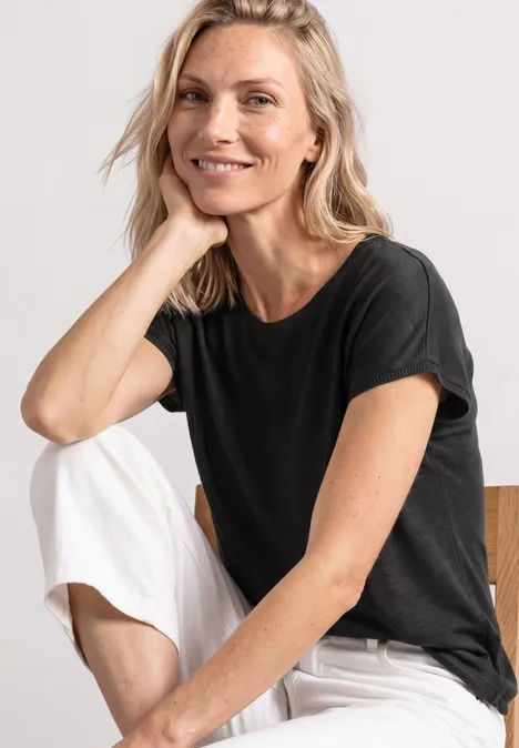 Dropped Shoulder Shirt mit Rundhals Black Frau sitzt in schwarzem T-Shirt und weißen Hosen vor neutralem Hintergrund.