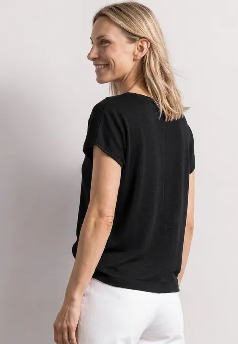 Dropped Shoulder Shirt mit Rundhals Black Frau im schwarzen T-Shirt und weißen Hosen, von hinten im Dreiviertelprofil vor neutralem Hintergrund.