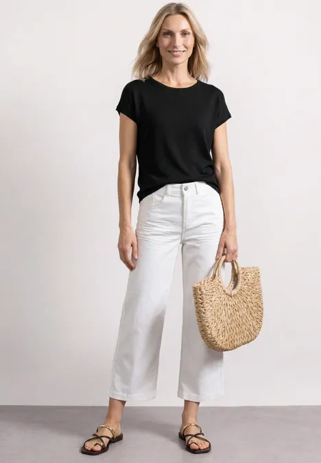 Dropped Shoulder Shirt mit Rundhals Black Frau in schwarzem T-Shirt, weißen Jeans und Sandalen trägt eine Strohtasche vor hellem Hintergrund.