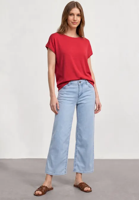 Vrouw in rood T-shirt, lichtblauwe wijdvallende jeans en bruine sandalen tegen neutrale achtergrond